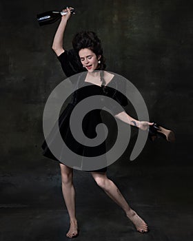 Full-length portrait of young beautiful happy woman in black holiday attire drinking wine and laughing isolated on dark