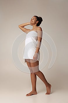 Full-length portrait of young beautiful woman posing in white bath towel isolated over gray studio background