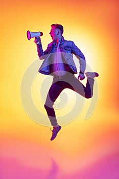 Full-length portrait of young excited man jumping with megaphone isolated on orange background in neon light, filter
