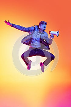 Full-length portrait of young excited man jumping with megaphone isolated on orange background in neon light, filter