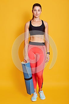 Full length portrait of young female with blue exercising mat in hand, posing isolated over yellow background, looking directlky