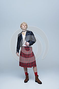 Full-length portrait of young handsome, curly haired man posing in Scottish kilt and leather jacket against grey studio