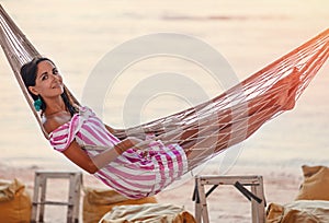 A full length portrait of young happy beautiful woman in a striped dress is lying on a wicker hammock.Side view.In the background,