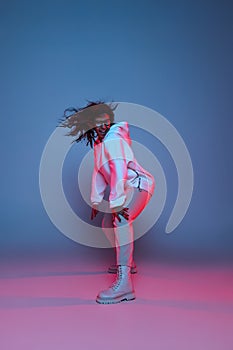 Full-length portrait of young happy dark skinned girl in hoodie jumping, dancing isolated on gradient blue-pink