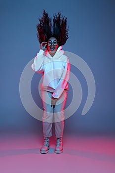 Full-length portrait of young happy dark skinned girl in hoodie jumping, dancing isolated on gradient blue-pink