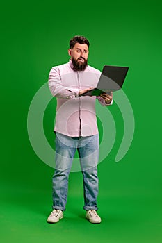 Full-length portrait of young man with beard looks at laptop screen with blank look against vibrant green studio