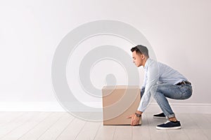 Full length portrait of young man lifting heavy cardboard box near white wall