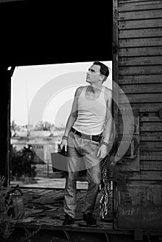 Full-length portrait of young man, wearing grey pants, white top, standing inside old railway carriage, leaning on doors. Black