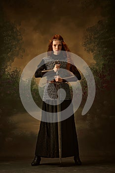 Full-length portrait of young princess in chainmail with sword posing against vintage background, showing her brave.