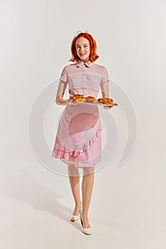 Full-length portrait of young redhead woman in cute pink dress with freshly baked buns  over grey background
