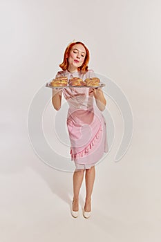 Full-length portrait of young redhead woman in cute pink dress serving freshly baked buns isolated over grey background