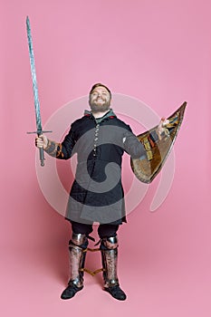 Full-length portrait of young smiling man, medieval warrior or archer in armor with sword and shield  over pink