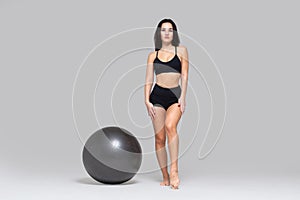 Full length portrait of young woman in black sportswear standing near fitness ball isolated on grey background
