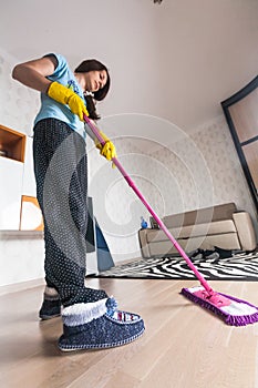 Full length portrait of young woman cleaning parquet floor in living room at home. Housewife with mop cleaning home