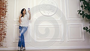 Full length portrait of young woman, girl, brunette, in white shirt and jeans, standing on background of white wall with