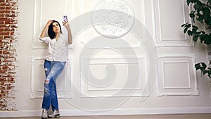 Full length portrait of young woman, girl, brunette, in white shirt and jeans, standing on background of white wall with