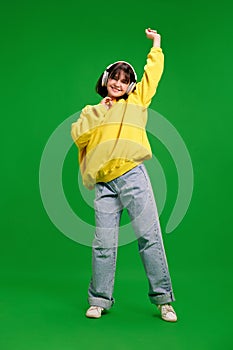 Full-length portrait of young woman in oversize outfit dancing while listening music in headphones against vibrant green