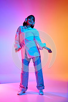 Full-length portrait of young woman in oversize outfit dancing while listening music in headphones in neon light against