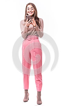 Full length portrait of a young woman typing a text message on her cell phone and leaning against a wall isolated on white
