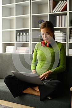 Full length of pretty young woman using laptop computer on couch at home. Freelance, distance, education, e-learning