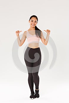 Full length of professional fitness coach, asian girl athelte pointing at herself and smiling happy, workout in gym