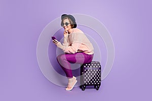Full length profile photo of pretty lady waiting registration flight hold telephone sitting on bag wear hat sun specs