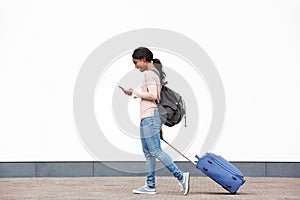 Full length profile young african american female traveler walking with cellphone and suitcase against white wall