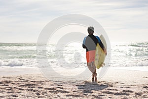 Full length rear view of biracial senior woman running with surfboard towards sea at sunny beach