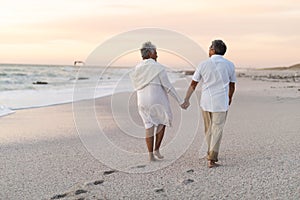 Full length rear view of senior multiracial couple holding hands walking at beach during sunset