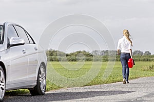 Full length rear view of woman carrying gas can leaving behind broken down car at countryside