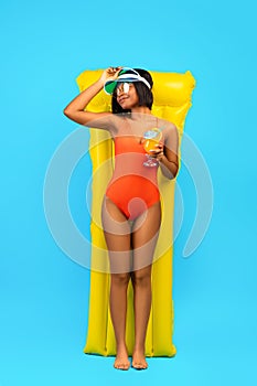 Full length of relaxed black lady in swimsuit lying on yellow inflatable lilo and drinking cocktail over blue background