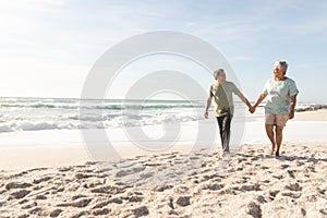 Full length of senior multiracial couple holding hands walking together at sunny beach against sky