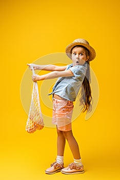 full length of shocked girl in straw hat holding reusable string bag with oranges on yellow.