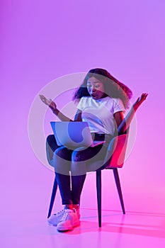 Full length of shocked young black woman using laptop, sitting in armchair, opening mouth in surprise in neon light