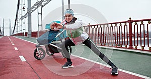 Full length shot of active young sportswoman stretching her body, standing on the bridge with a baby carriage, preparing