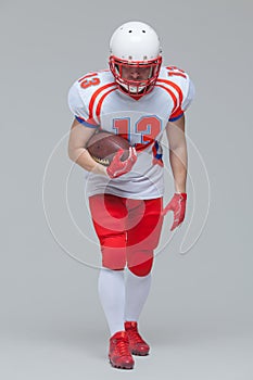 Full length shot of american football sportsman player wearing helmet posing with rugby ball isolated on grey background