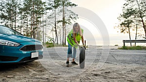 Full length shot of attractive young woman wearing reflective vest, rolling spare tire to change the flat one on the