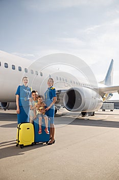 Full length shot of beautiful stewardesses standing near two kids sitting on their luggage in front of big airplane and