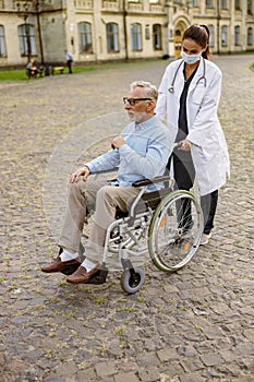 Full length shot of caring young nurse in protective mask taking care of senior man handicapped patient in wheelchair