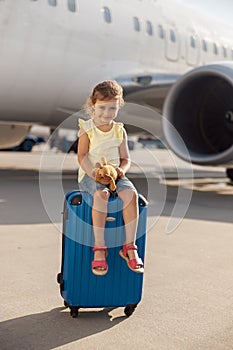 Full length shot of cute little girl holding her toy, looking at camera and sitting on suitcase in front of big airplane