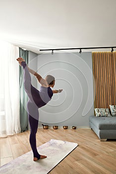 Full length shot of flexible young woman exercising, doing Lord of the dance yoga pose while standing on a mat in modern