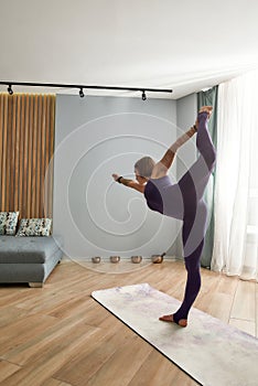 Full length shot of flexible young woman in purple bodysuit exercising, doing Lord of the dance yoga pose while standing