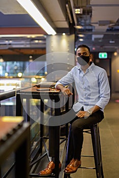 Full length shot of of handsome black African businessman sitting inside shopping mall while wearing face mask to