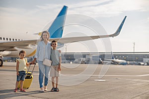 Full length shot of happy mother with two little kids smiling at camera while standing in front of big airplane in the