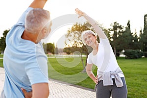 Full length shot of happy smiling mature family man and woman in sportswear stretching arms while warming up together outdoors in