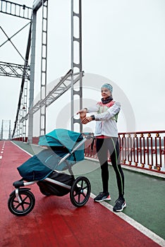 Full length shot of motivated young woman stretching her hands, standing on the bridge with a baby carriage on a cloudy