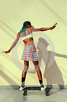 Full-length shot of a skater woman`s back wearing fancy clothes and riding her skateboard in front of a yellow wall with shadows photo