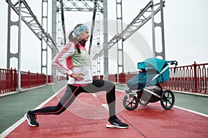 Full length shot of sportive woman stretching her legs, standing on the bridge with a baby carriage, preparing for a jog