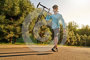 Full length shot of strong athletic male cyclist in sportswear and protective helmet carrying his bike after training in