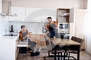 Full length shot of two handymen, workers in uniform talking while assembling kitchen cabinet using screwdriver indoors